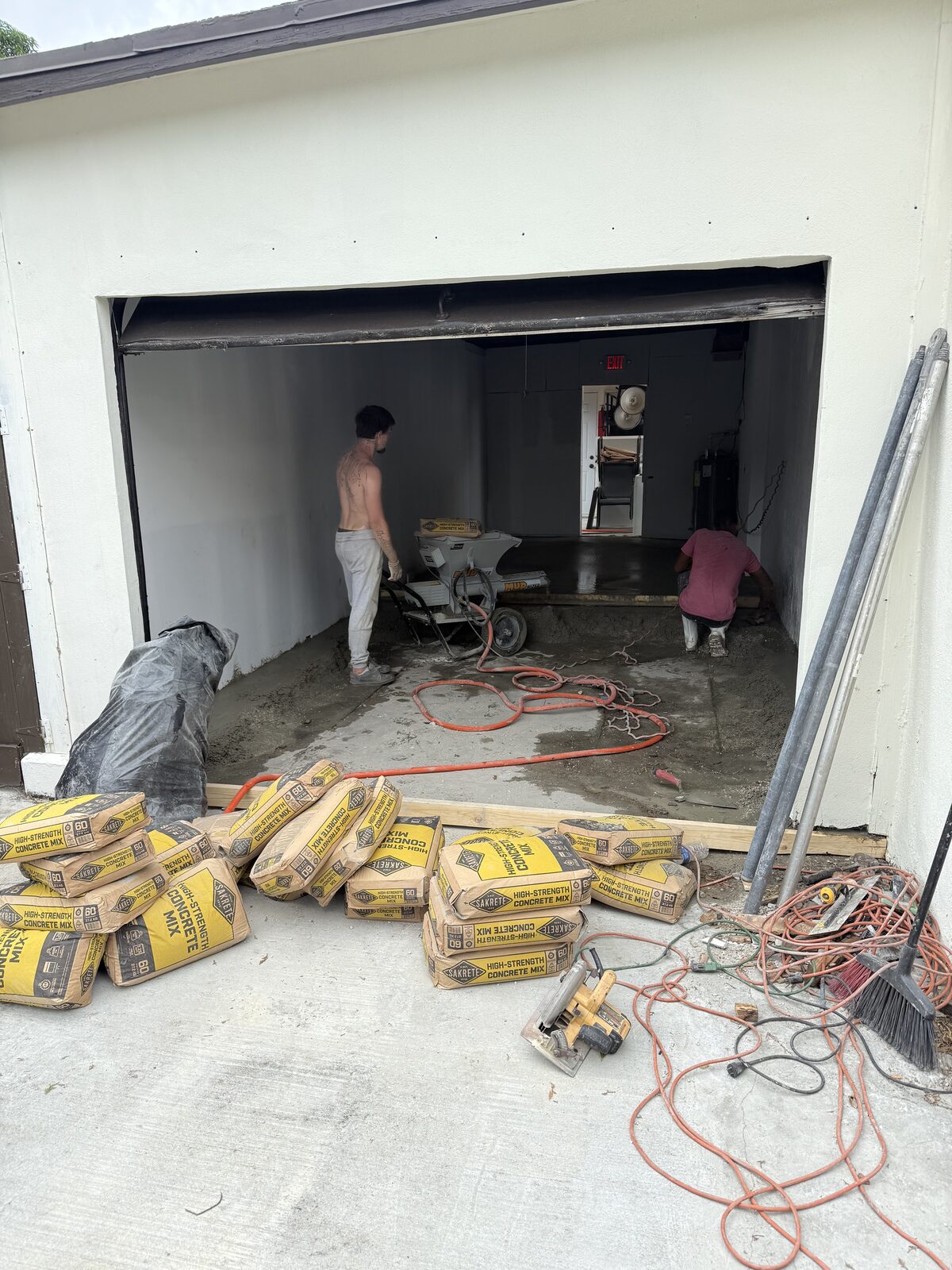 Bags of Sakrete high-strength concrete mix stacked outside the open garage door with crew working inside the bay beyond