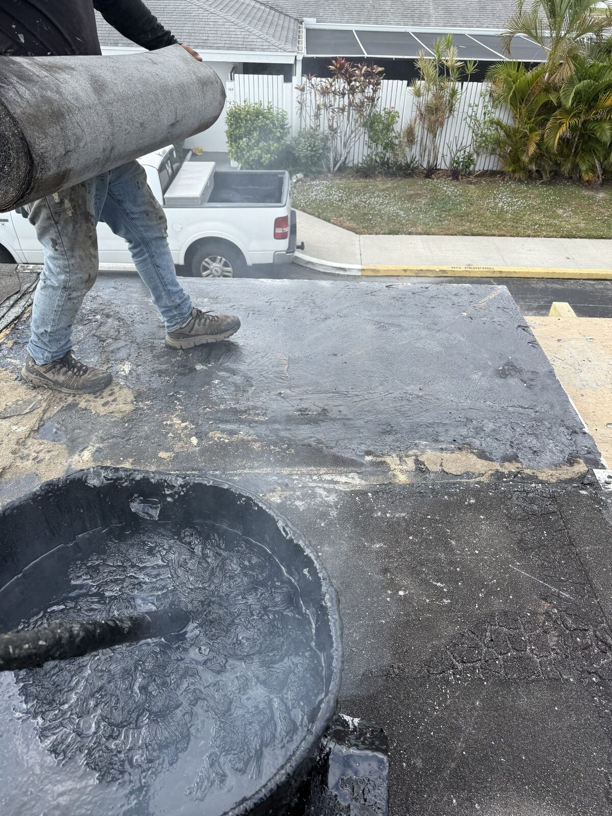 Roofer carrying a roll of modified-bitumen membrane across the carport deck beside a steaming bucket of hot asphalt