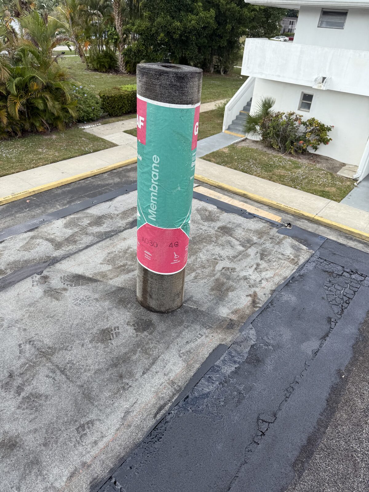 Roll of GAF modified-bitumen membrane standing upright on the prepared carport deck, the community building behind