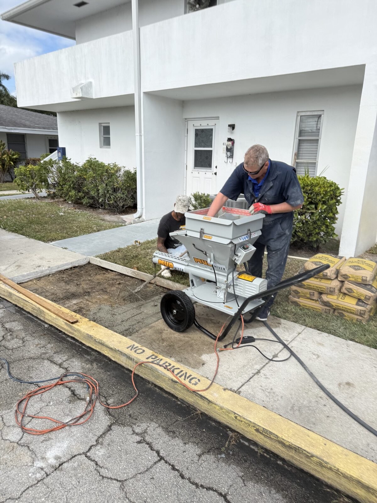 Worker feeding a bag of high-strength concrete mix into a continuous mixer beside the open sidewalk bay, stacked bags alongside
