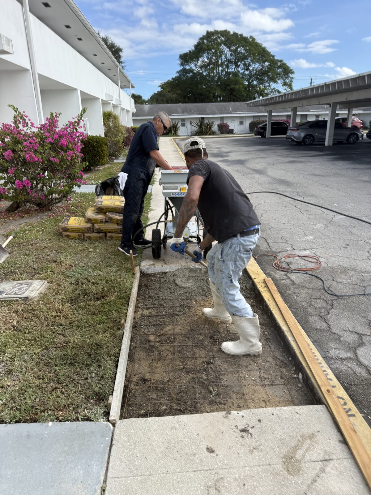 Two-man crew placing fresh concrete into the formed sidewalk bay over welded wire mesh outside a Royal Palm Beach villa building