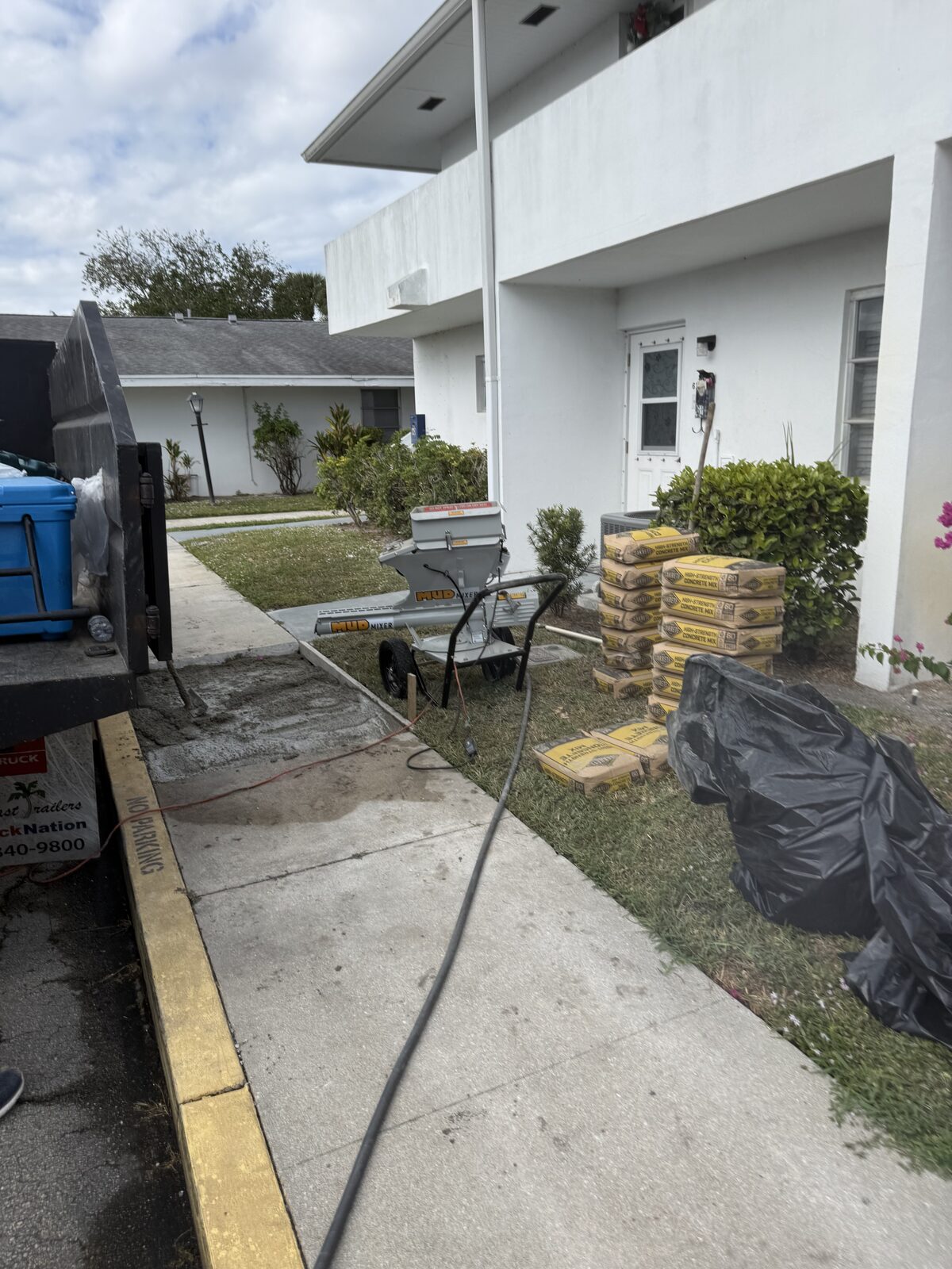Section of failed sidewalk removed from a walkway outside villa units, with a continuous concrete mixer staged and pallets of bagged high-strength concrete mix waiting on the grass