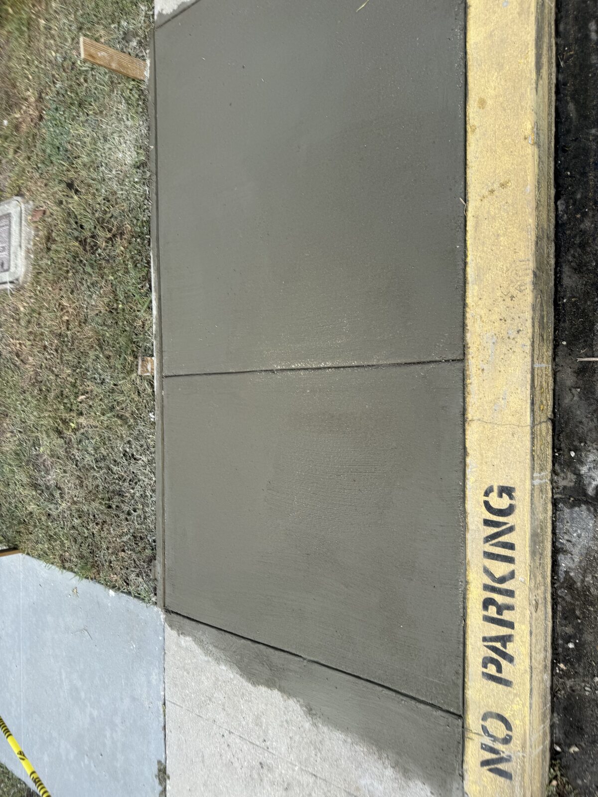 Two freshly poured and finished concrete walkway panels with a control joint, beside a yellow painted No Parking curb