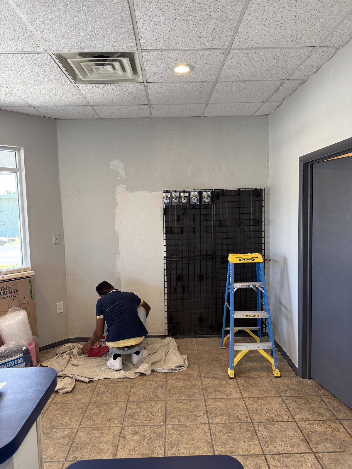 Painter patching a lobby wall beside the merchandising grid while the storage office stays open — Bell Engineering & Construction