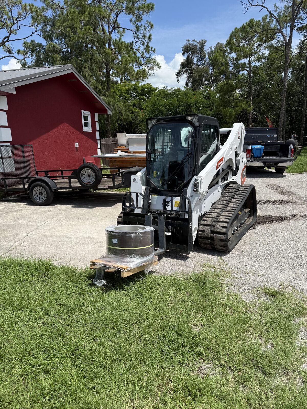 Bobcat compact track loader with pallet forks carrying a pallet of gutter coil stock across a job yard in Palm Beach County