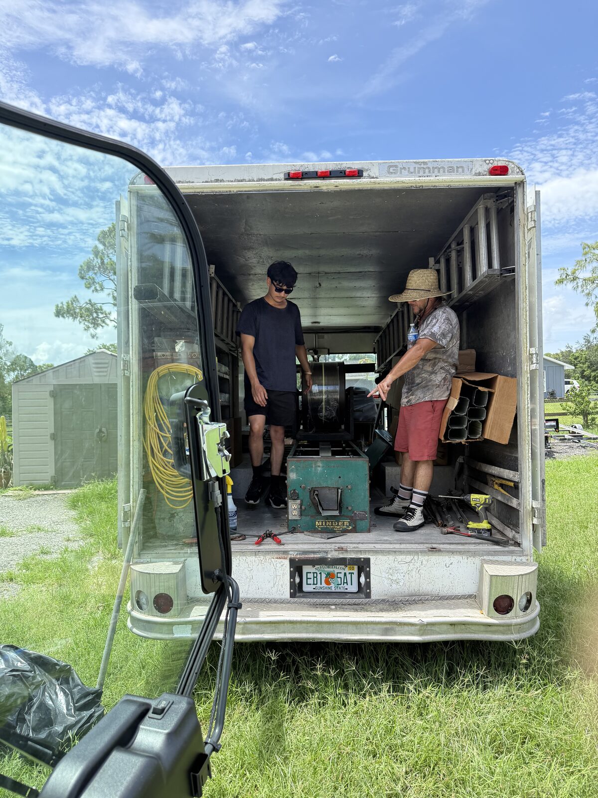 Two crew members working at the open rear of a Grumman step van fitted with a seamless gutter roll-forming machine and coil reel