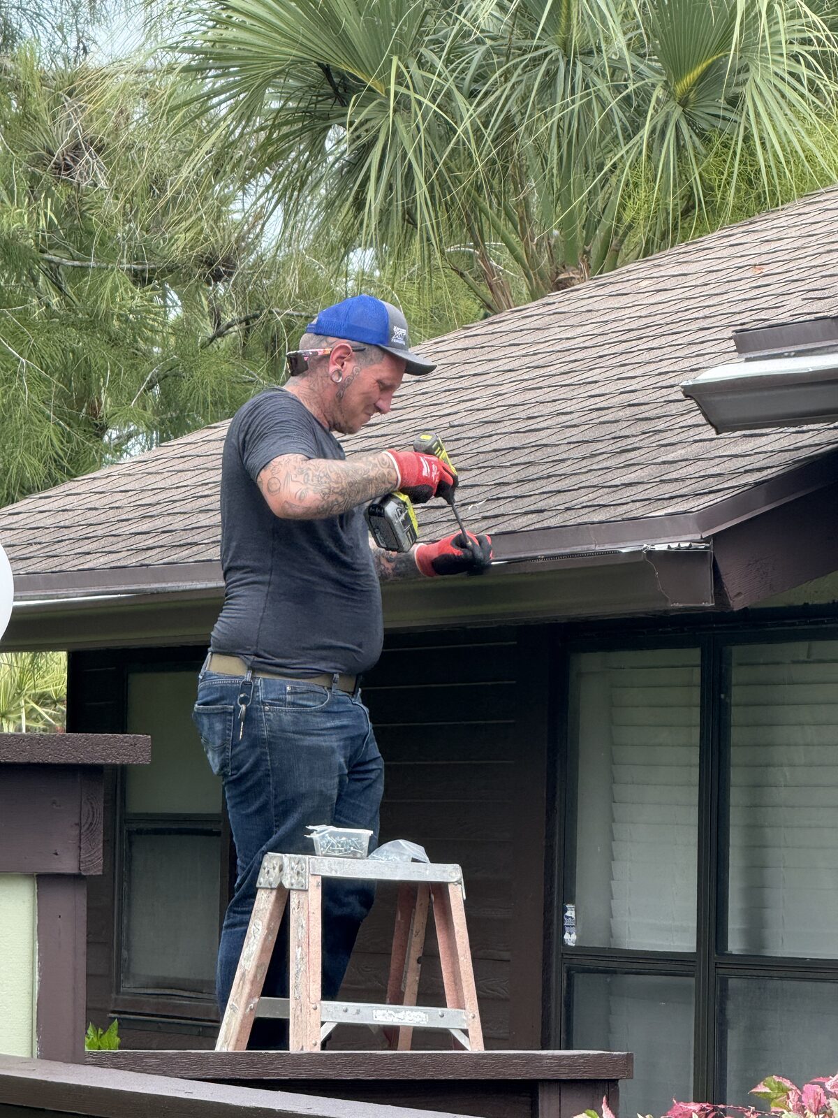Installer on a stepladder fastening a hidden hanger through a new brown seamless gutter at the eave of a shingle roof