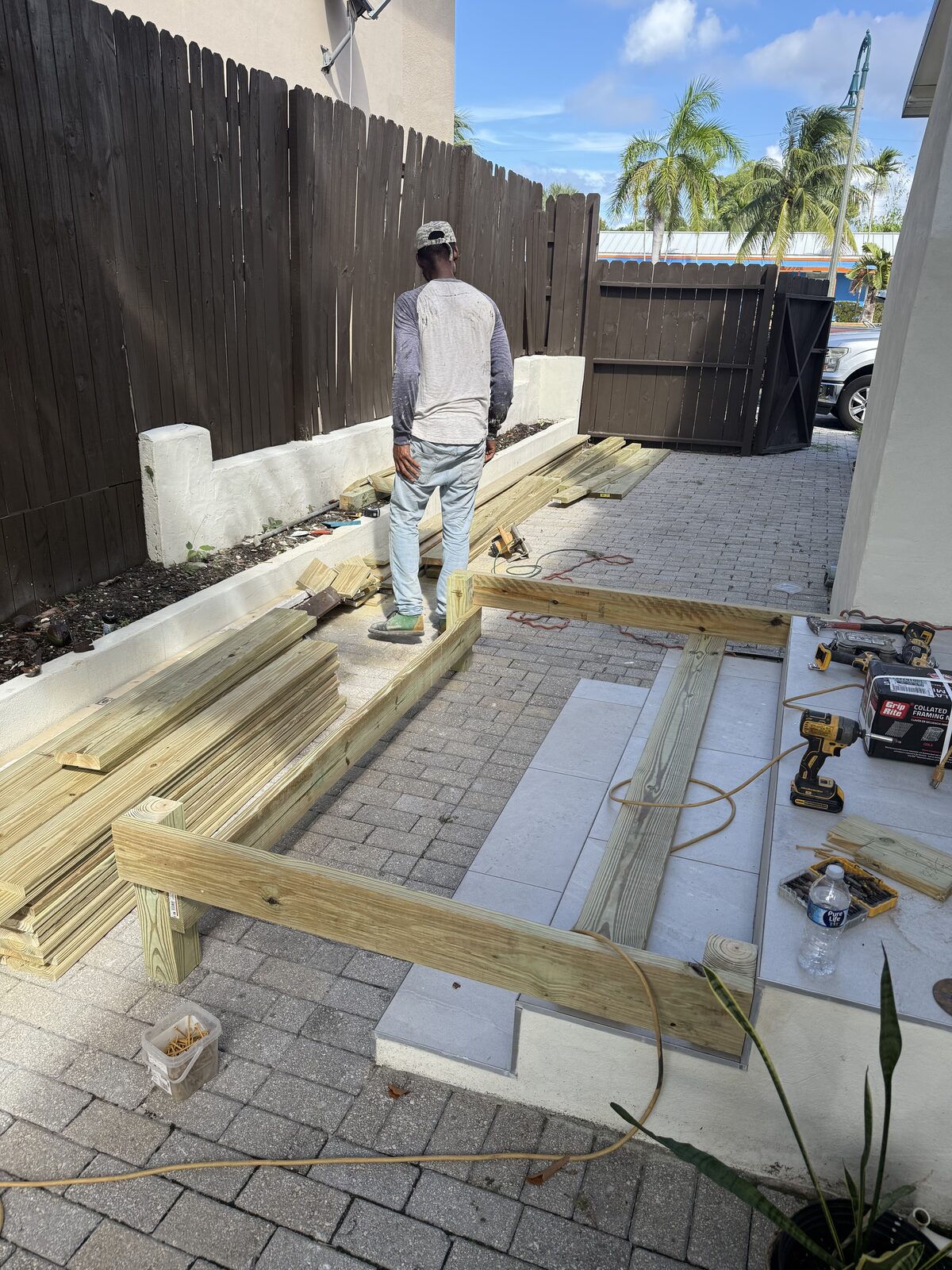 Pressure-treated beams and posts laid out on paver patio beside a stucco house, with framing nailer, collated nails and cordless drill staged on the tiled entry landing