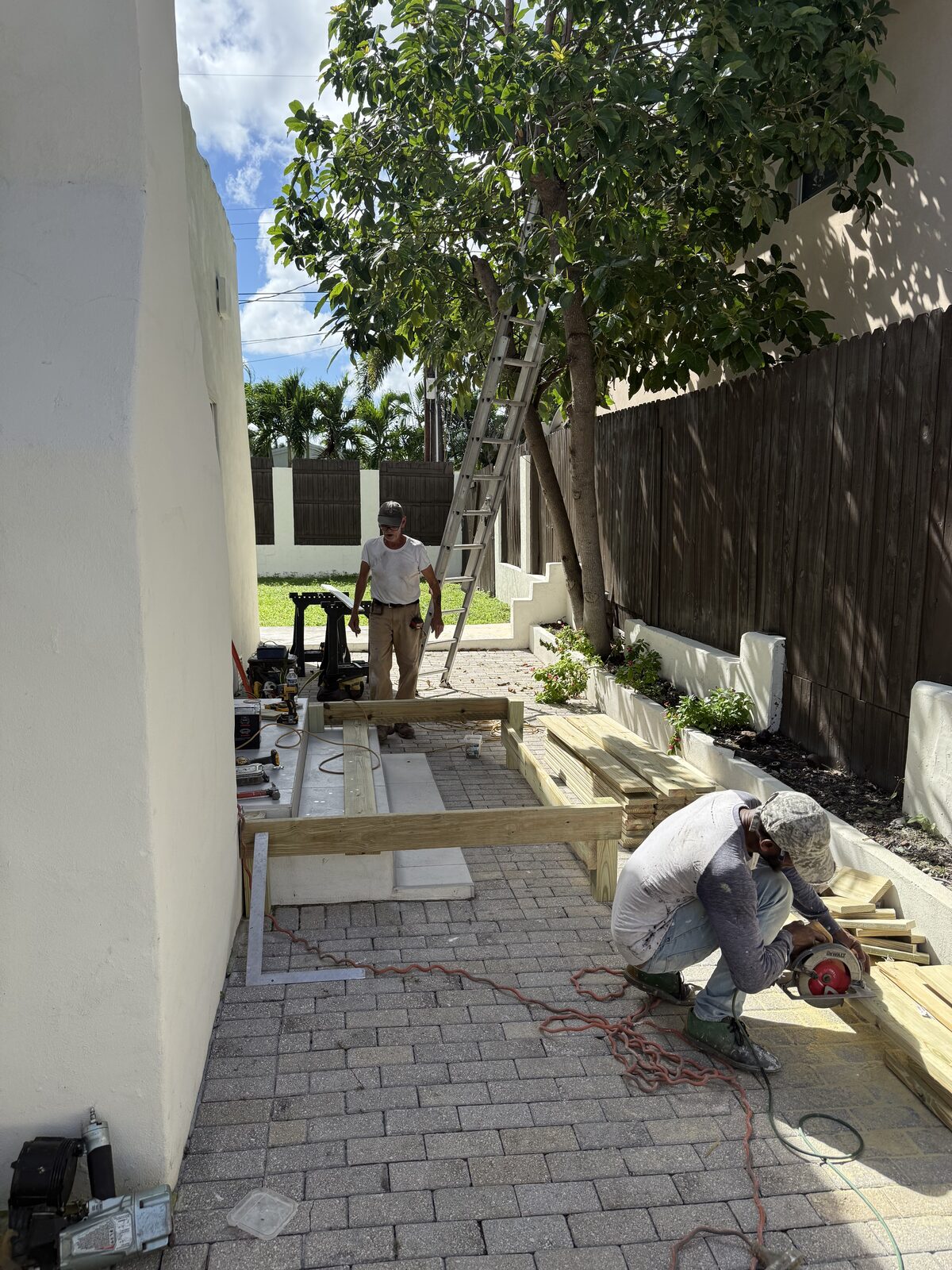 Two crew members working in the narrow side yard, one cutting pressure-treated lumber with a circular saw while the perimeter frame sits in place on the pavers