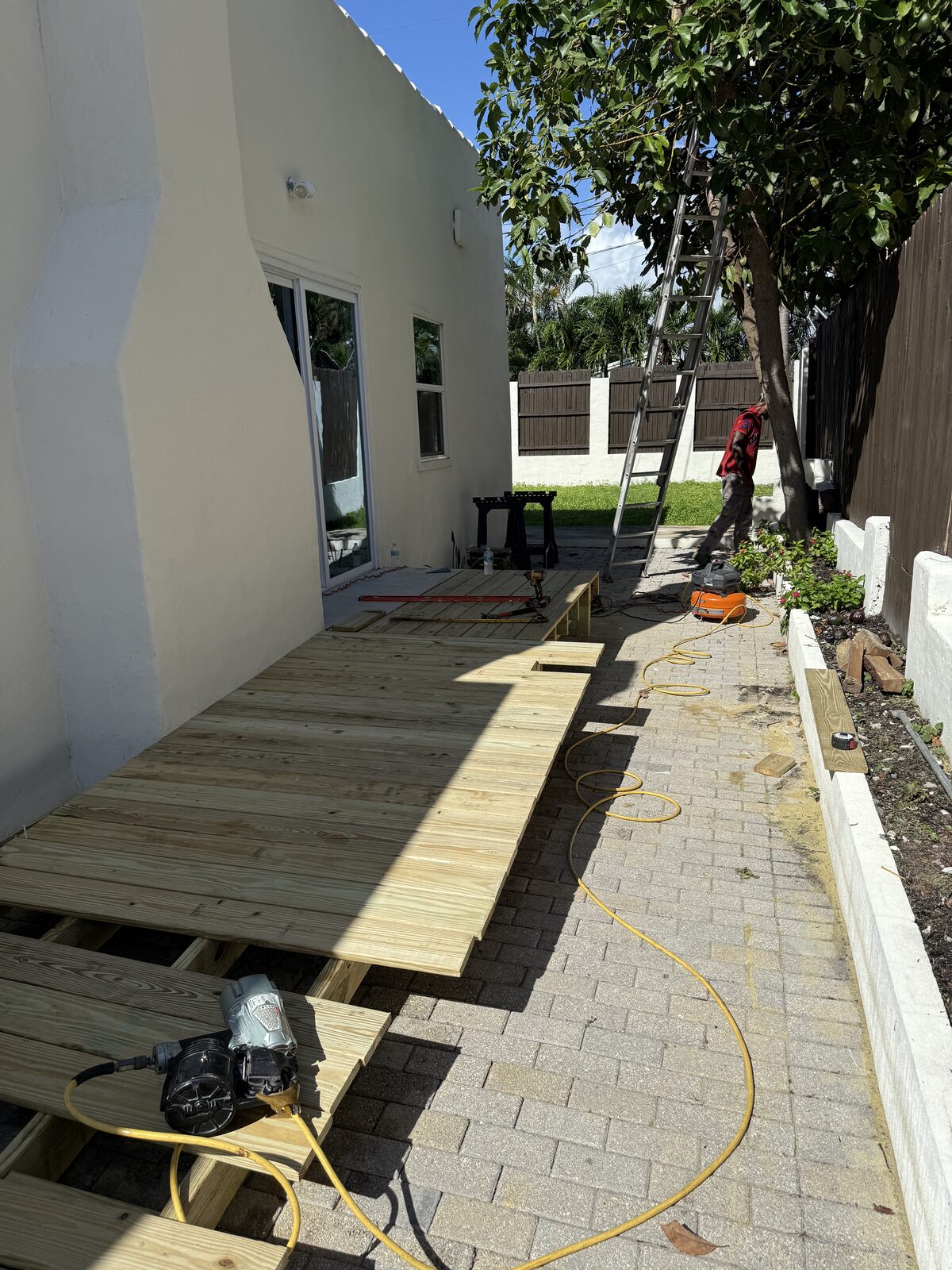 Deck boards being fixed across the ramp frame beside the stucco wall, pneumatic framing nailer and compressor hose on the finished section