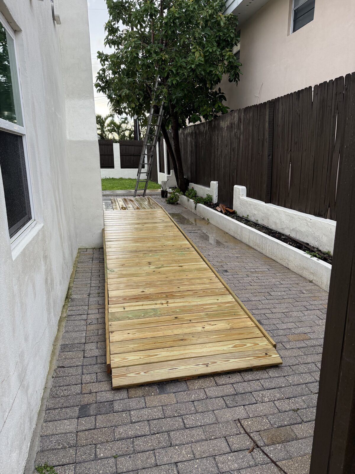 Completed timber ramp photographed after rain, water standing on the pavers alongside while the deck surface drains through the gaps between boards