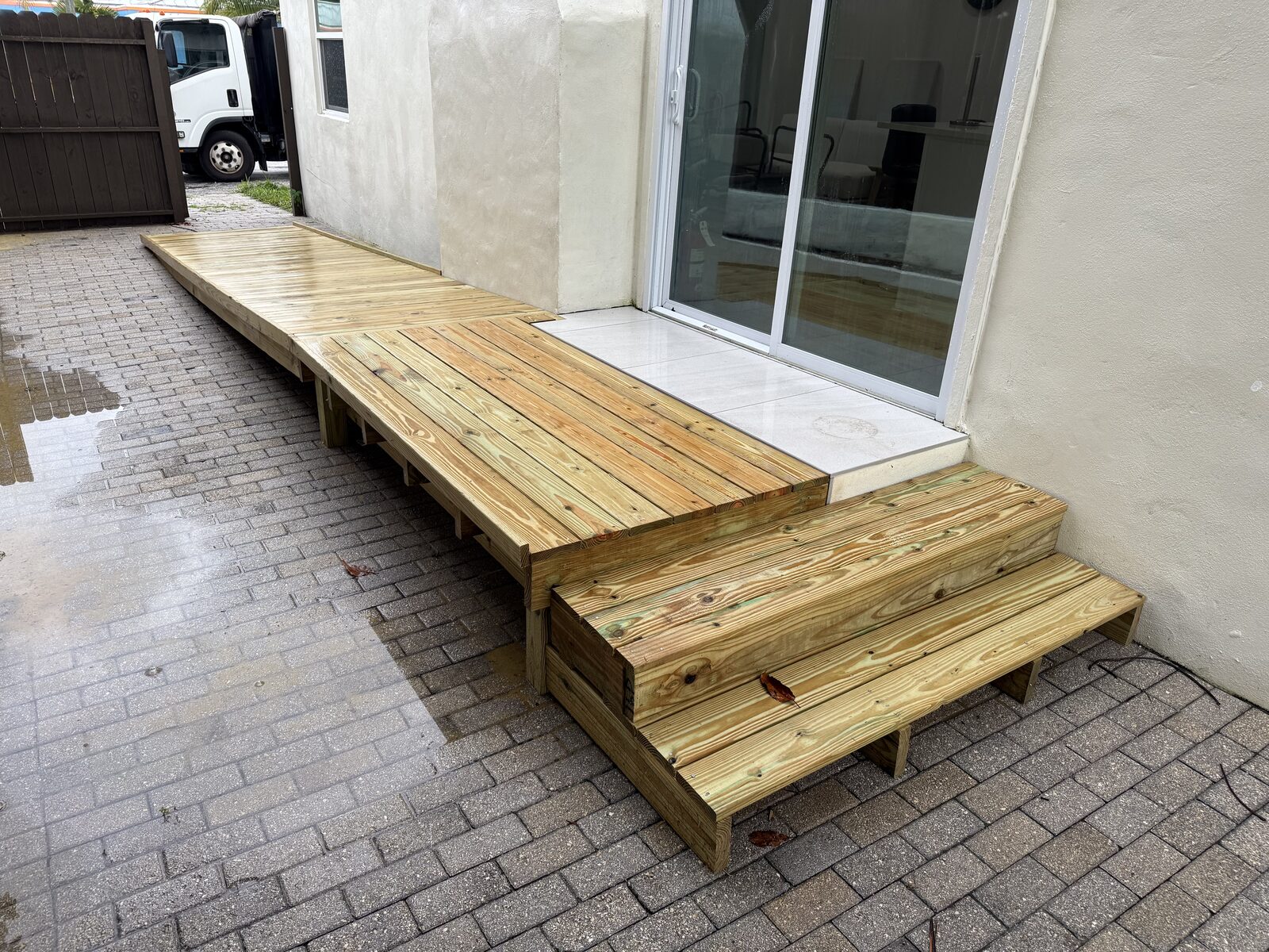 Finished pressure-treated wheelchair ramp running out along a paver side yard to a landing built flush with a sliding-glass door threshold, with two timber steps alongside