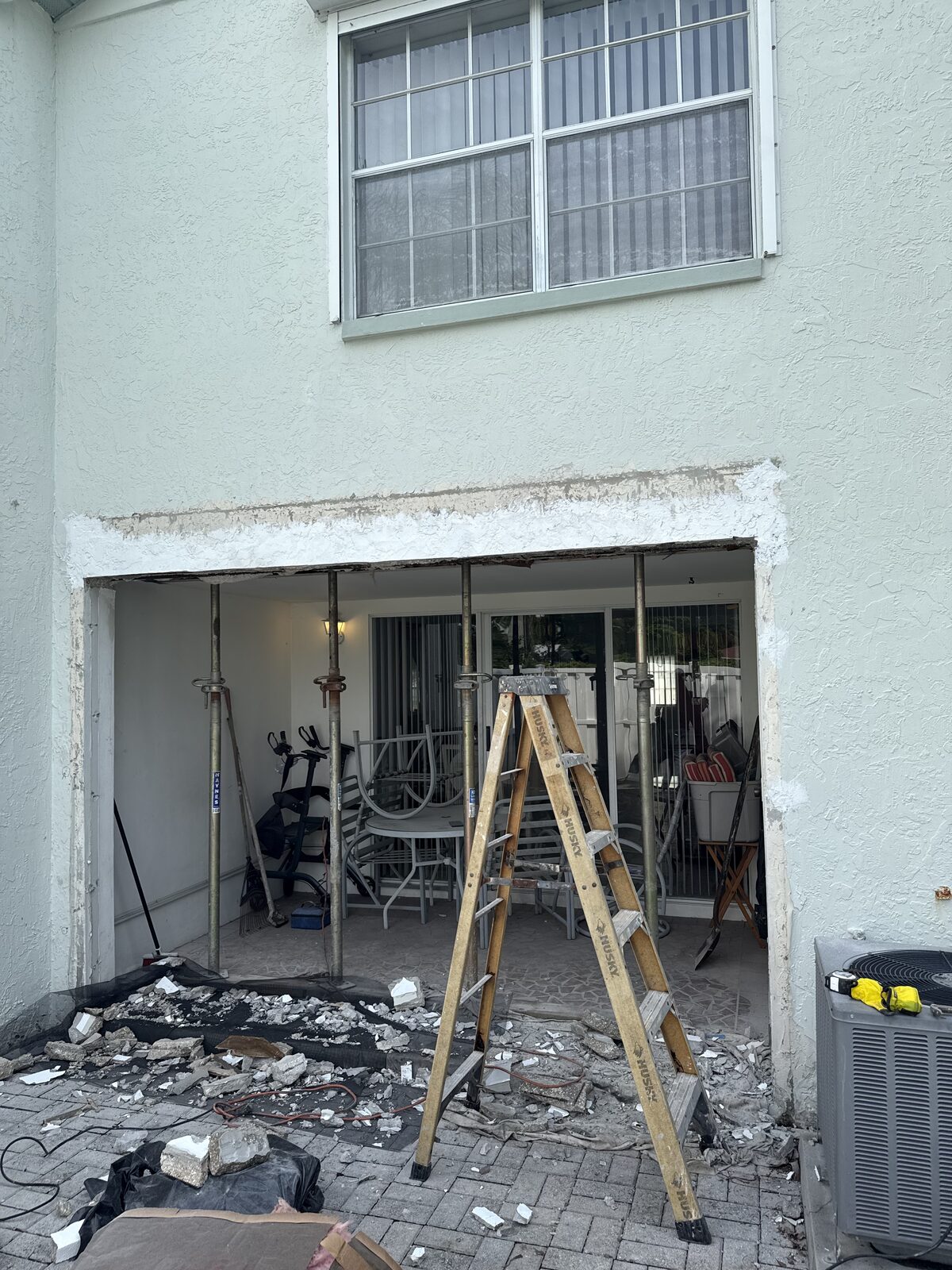 Wide view of a two-storey stucco home's covered patio opening with adjustable steel shoring posts installed across it and demolition debris on plastic sheeting below
