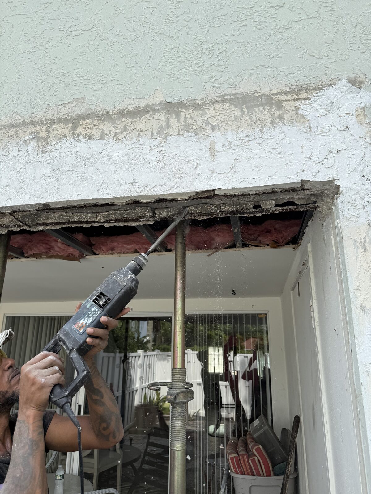 Worker using a rotary hammer overhead to break failed concrete out of a beam above a patio opening, insulation and framing visible in the cavity behind