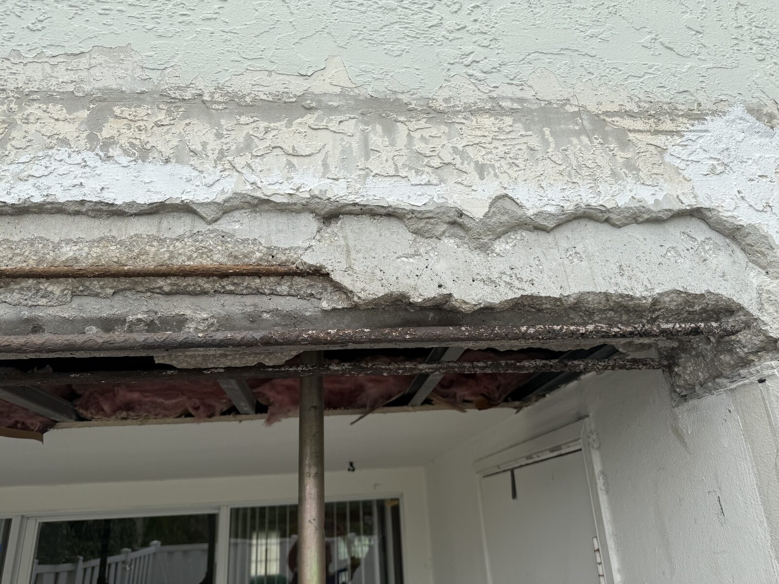 Spalled concrete beam above a patio opening with stucco peeled back in a horizontal band and corroded reinforcing bar exposed along its length