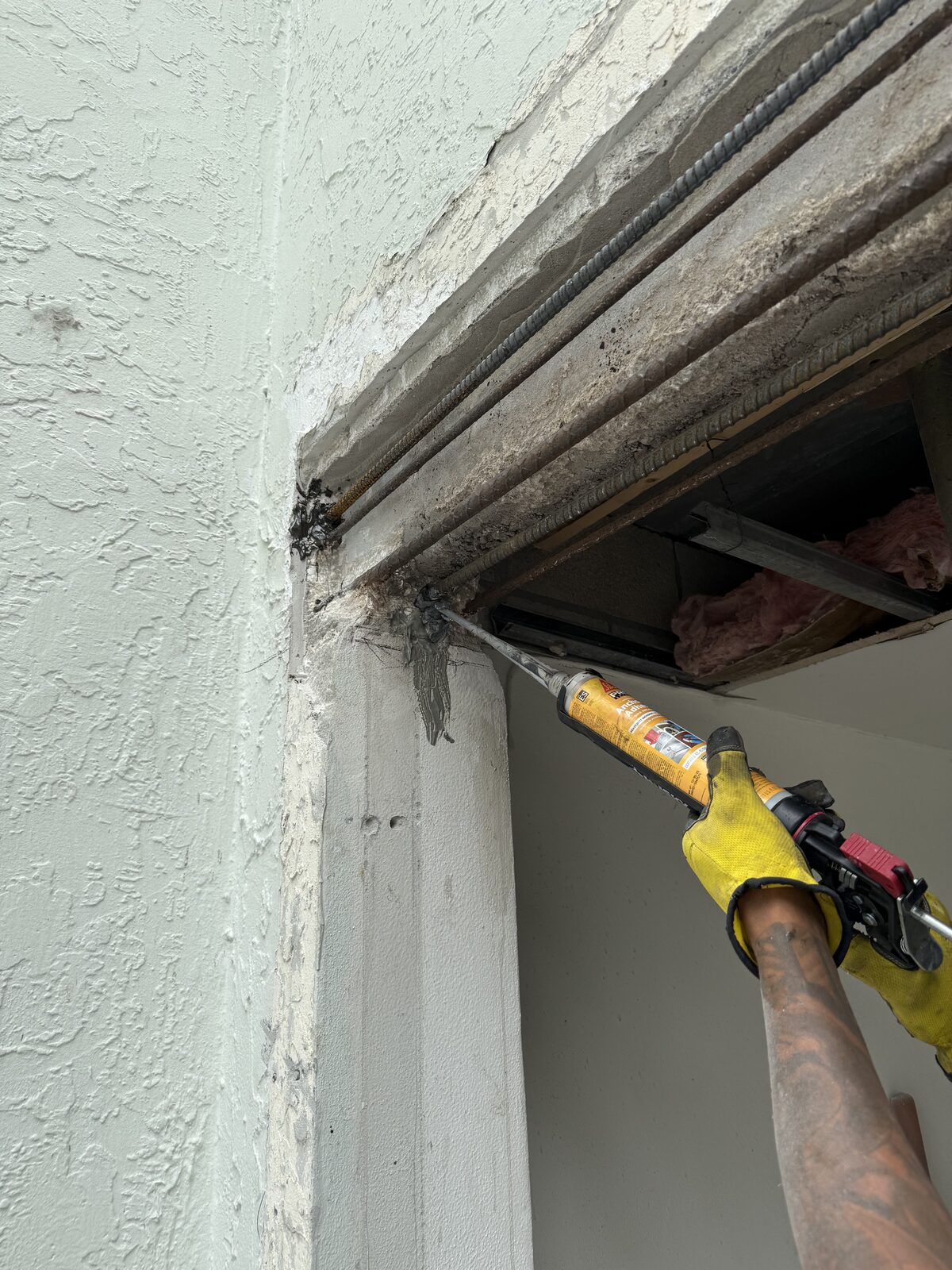 Gloved hand gunning anchoring adhesive into the pocket where the restored beam bears on the existing wall, new reinforcing bar in place alongside