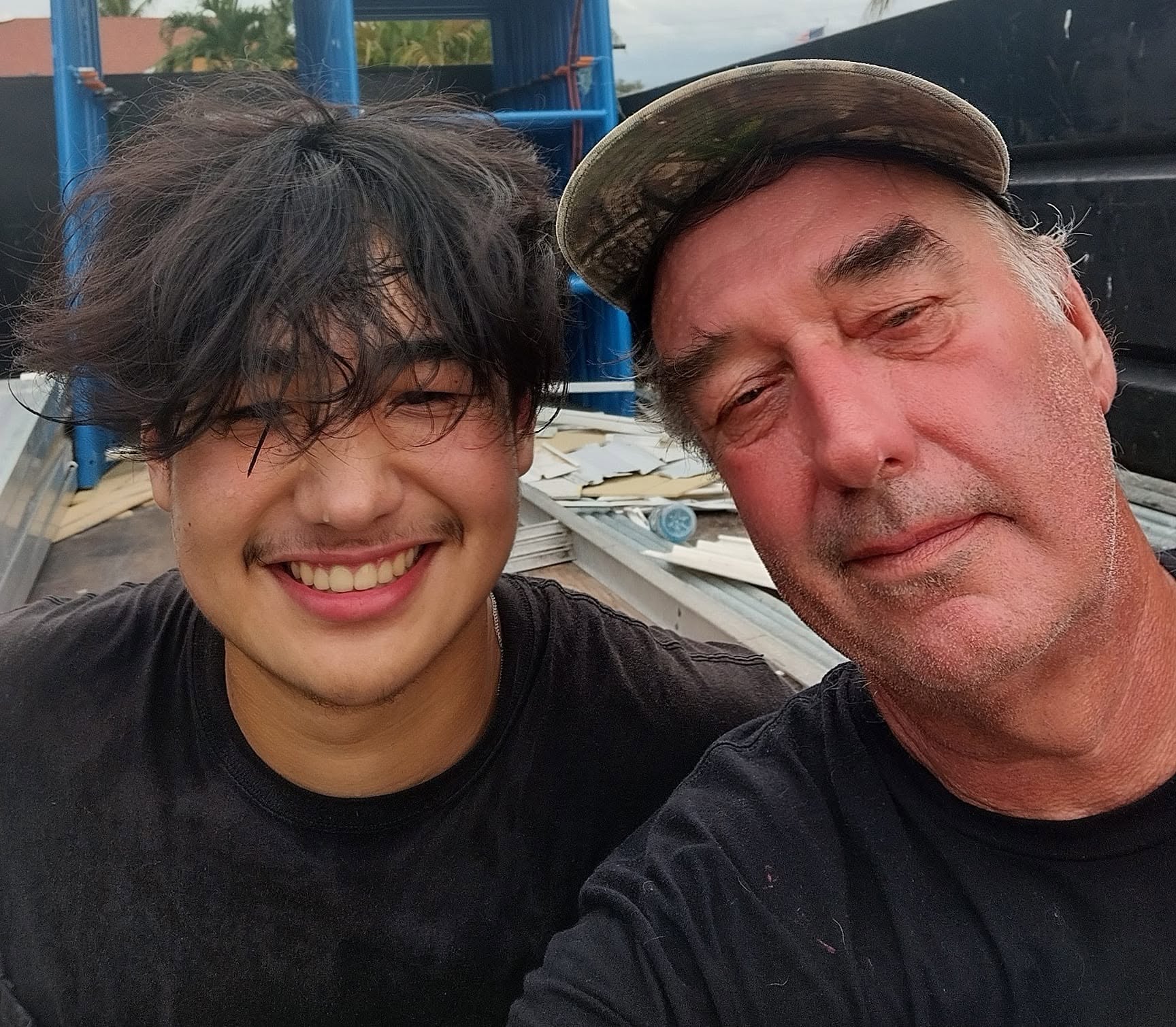 Randolph Scott Bell and his son Buddy on a roof in Fort Myers after Hurricane Ian, torn-off roofing panels and debris stacked behind them