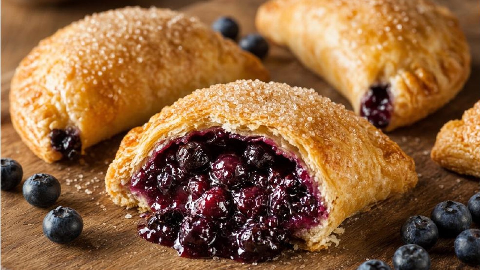 Florida Blueberry Hand Pie — a golden, turbinado-sugar-crusted pastry split open to show the bubbling blueberry filling, with two whole hand pies and fresh blueberries on a wooden board