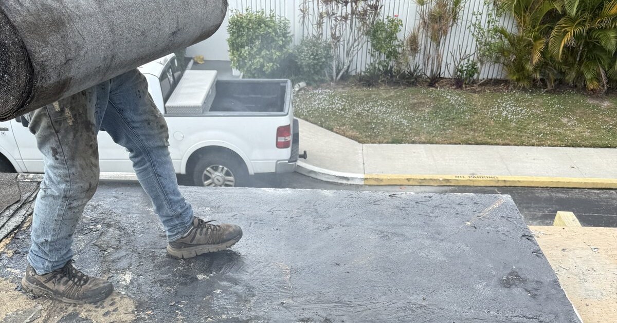 A crew member carries a roll of modified bitumen membrane across a common-element flat roof in a Palm Beach County HOA community, with the community's parking, fence line and palms behind