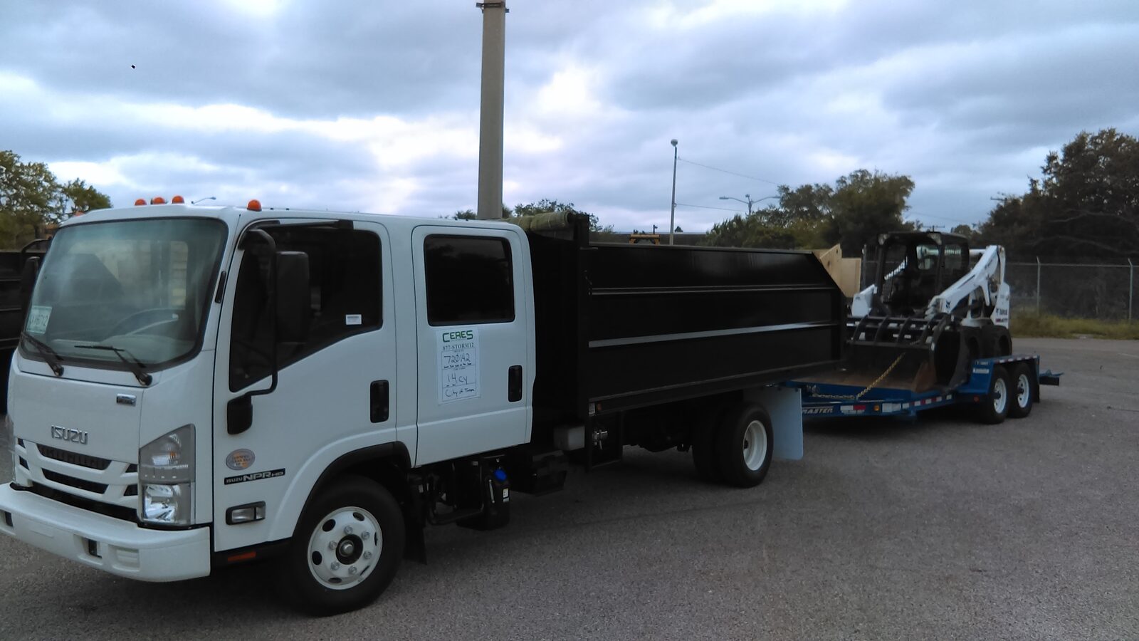 Isuzu NPR-HD crew cab dump truck towing a lowboy trailer carrying a Bobcat skid steer with a grapple attachment
