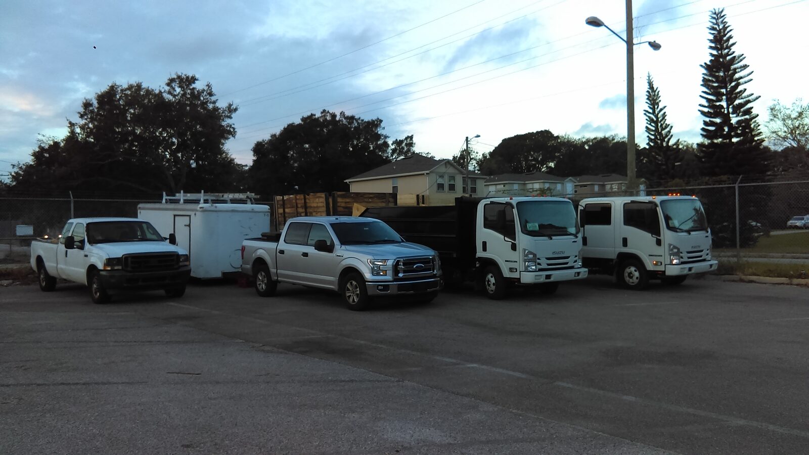 Storm-cleanup fleet lined up at dusk in a Tampa staging lot — pickup trucks, an enclosed trailer and two Isuzu cabover dump trucks