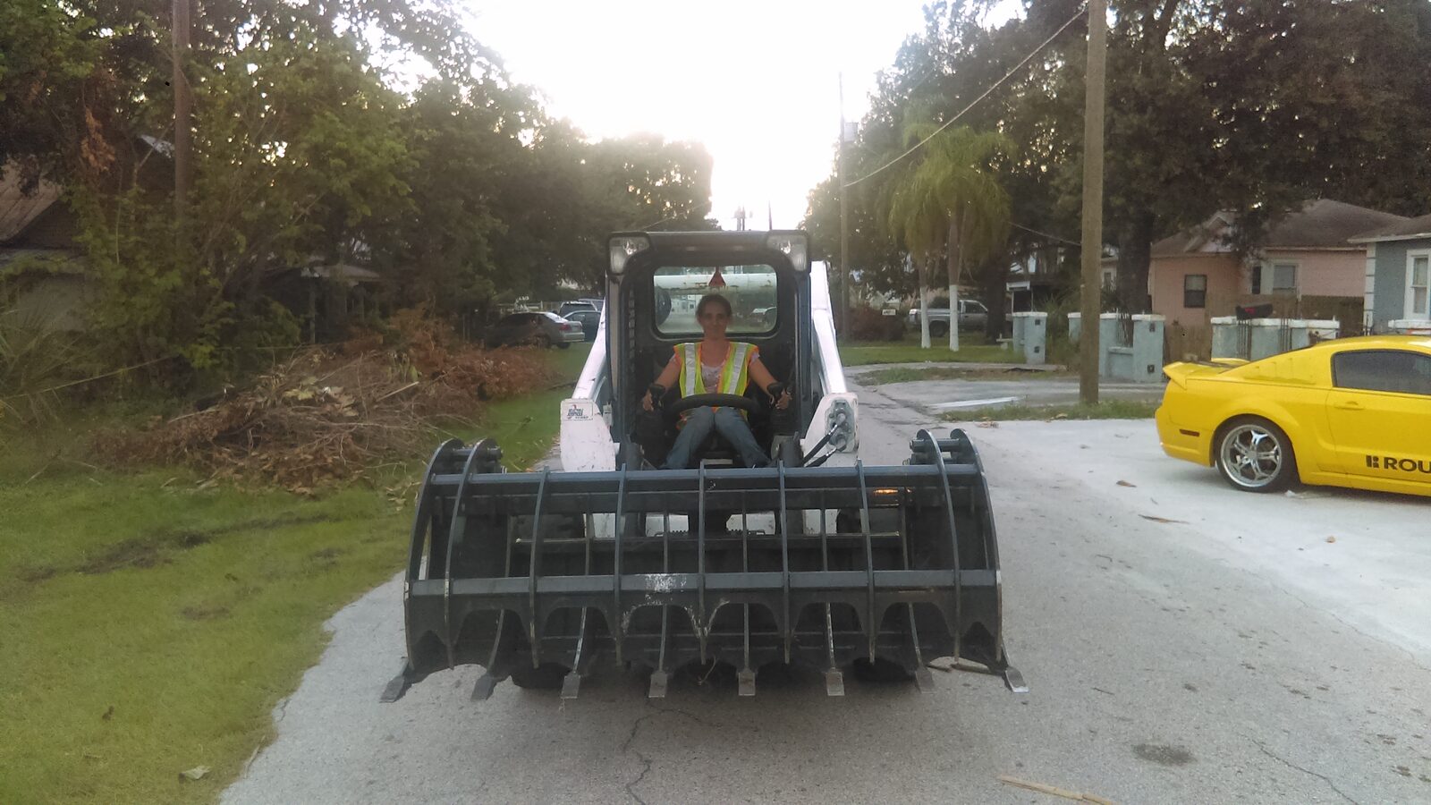 Operator in a hi-vis vest seated in a Bobcat skid steer fitted with a wide root grapple on a residential Tampa street